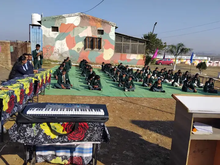 Students in blazers sit in rows on the ground for an outdoor school programme.