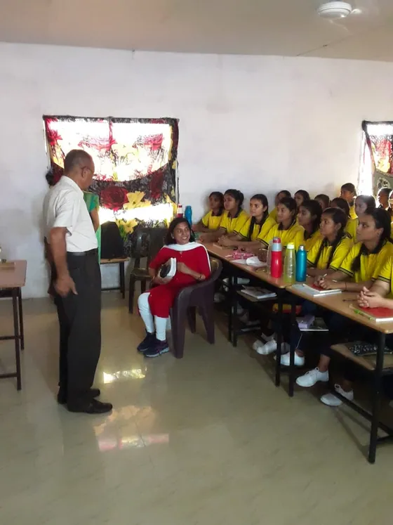 A teacher addresses girl students seated at long desks in a classroom.