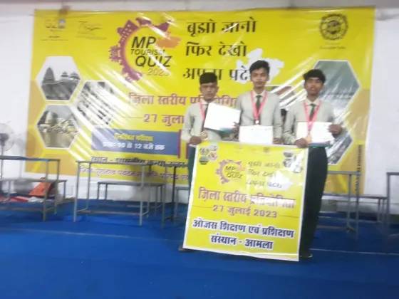 Three students in school uniform hold medals and certificates beside an Ojas banner at the MP Tourism Quiz 2023.