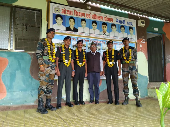 Six young men in Army uniform, garlanded with marigolds, stand with a mentor in front of the school's meritorious-students board.