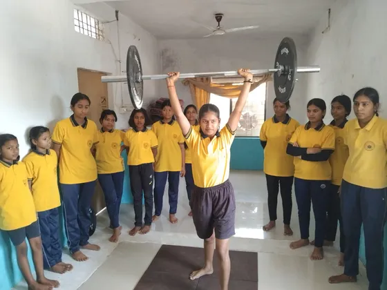 A student lifts a barbell overhead in the training hall as her classmates look on.
