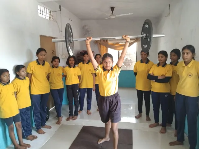 A student lifts a barbell overhead in the training hall as her classmates look on.