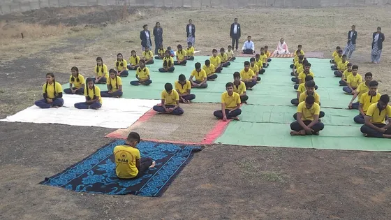 Rows of students sit cross-legged on mats in an outdoor yoga session on the campus ground.