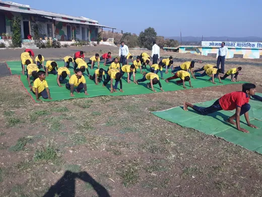 Students in yellow T-shirts perform surya namaskar on green mats on the campus ground.